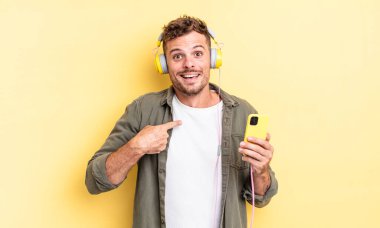 Genç yakışıklı adam mutlu hissediyor ve heyecanlı kulaklıklar ve akıllı telefon konseptiyle kendini işaret ediyor.
