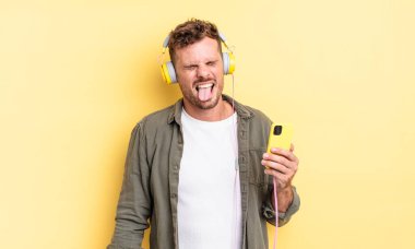 Neşeli ve asi tavırları olan genç yakışıklı bir adam, şaka yapıyor, kulaklık takıyor ve akıllı telefon konsepti kullanıyor.