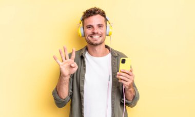 Genç yakışıklı adam gülümsüyor ve arkadaş canlısı görünüyor. Dört numaralı kulaklığı ve akıllı telefon konseptini gösteriyor.