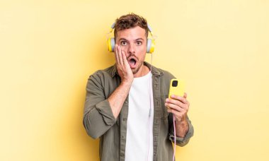 Genç yakışıklı adam şok ve korkmuş kulaklıklar ve akıllı telefon konsepti hissediyor.