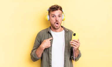 Genç ve yakışıklı bir adam ağzı açık, kulaklıkları ve akıllı telefon konseptini işaret ederek şok olmuş ve şaşırmış görünüyor.