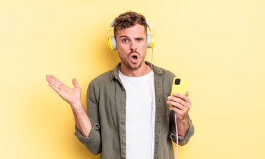 Genç yakışıklı adam şaşırmış ve şok olmuş görünüyor, çenesi düşmüş, elinde bir nesne kulaklığı ve akıllı telefon konseptiyle.
