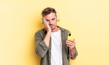 Genç yakışıklı bir adam yorgun kulaklıklar ve akıllı telefon konseptinden sonra sıkılmış, hayal kırıklığına uğramış ve uykulu hissediyor.