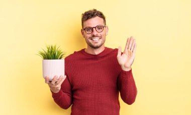 Genç yakışıklı adam mutlu bir şekilde gülümsüyor, el sallıyor, sizi selamlıyor ve selamlıyor. dekoratif bitki konsepti