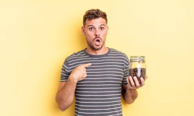 Genç ve yakışıklı bir adam ağzı açık, şaşırmış ve şaşırmış görünüyor. Kahve çekirdekleri kavramına işaret ediyor.