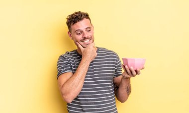 Genç ve yakışıklı bir adam mutlu, kendinden emin bir ifadeyle gülümsüyor ve elleri çenesinde boş kase konseptiyle.