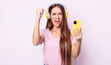 Genç ve güzel bir kadın sinirli bir ifadeyle agresif bir şekilde bağırıyor. Kulaklık ve akıllı telefon.