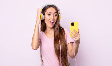 Genç ve güzel bir kadın kendini mutlu, heyecanlı ve şaşırmış hissediyor. Kulaklık ve akıllı telefon.