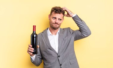 Kafası karışmış, kafası karışmış, kaşınan genç, yakışıklı bir adam. Şarap şişesi konsepti