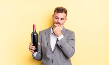 Genç ve yakışıklı bir adam mutlu ve kendinden emin bir ifadeyle gülümsüyor, elleri çenesinde. Şarap şişesi konsepti