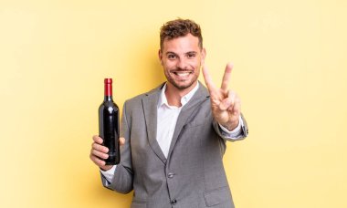 Genç yakışıklı adam gülümsüyor ve mutlu görünüyor, zafer ya da barış işareti yapıyor. Şarap şişesi konsepti