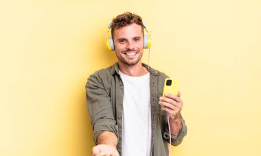 Genç yakışıklı adam mutlu bir şekilde gülümsüyor ve bir konsept kulaklık ve akıllı telefon konsepti sunuyor.