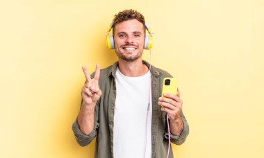 Genç yakışıklı adam gülümsüyor ve arkadaş canlısı görünüyor. İki numaralı kulaklığı ve akıllı telefon konseptini gösteriyor.
