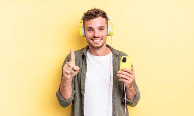 Genç yakışıklı adam gülümsüyor ve arkadaş canlısı görünüyor. Bir numaralı kulaklıkları ve akıllı telefon konseptini gösteriyor.