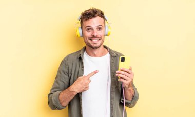 Genç yakışıklı adam neşeyle gülümsüyor, mutlu hissediyor ve kulaklıkları ve akıllı telefon konseptini işaret ediyor.