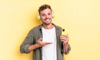 Genç ve yakışıklı adam neşeyle gülümsüyor, mutlu hissediyor ve bir konsept kulaklık ve akıllı telefon konsepti gösteriyor.