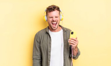 Genç yakışıklı adam agresif bir şekilde bağırıyor, çok kızgın kulaklıklar ve akıllı telefon konsepti takıyor.