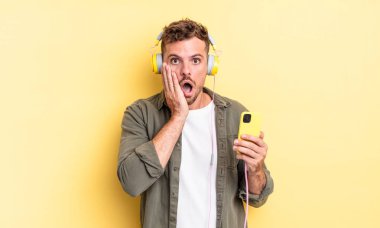 Genç yakışıklı adam şok ve korkmuş kulaklıklar ve akıllı telefon konsepti hissediyor.