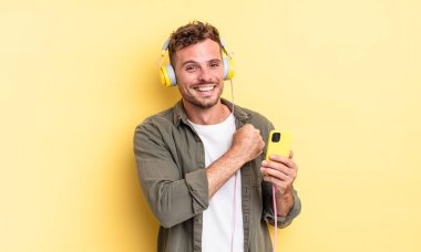 Genç yakışıklı adam kendini mutlu hissediyor ve bir meydan okumayla karşı karşıya ya da kulaklık ve akıllı telefon konseptini kutluyor.