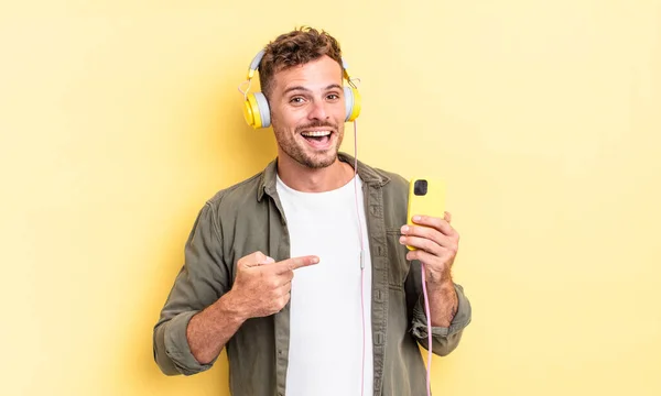 Mladý Pohledný Muž Vypadá Vzrušeně Překvapeně Ukazuje Boční Sluchátka Smartphone — Stock fotografie