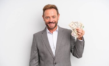 Orta yaşlı yakışıklı adam mutlu bir şekilde gülümsüyor, eli kalçasında ve kendinden emin. Dolar banknotları konsepti