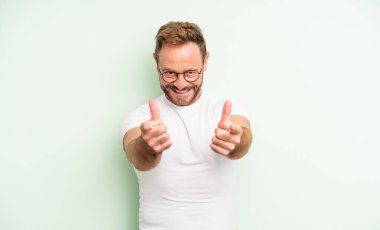 Orta yaşlı, yakışıklı, pozitif, başarılı, mutlu bir tavırla gülümseyen, kamerayı işaret eden, elleriyle silah işareti yapan bir adam.