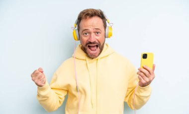 Orta yaşlı yakışıklı adam şokta, gülüyor ve başarıyı kutluyor. kulaklıklar ve akıllı telefon konsepti