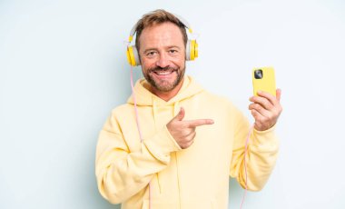 Orta yaşlı yakışıklı bir adam neşeli bir şekilde gülümsüyor, mutlu hissediyor ve bir tarafı işaret ediyor. kulaklıklar ve akıllı telefon konsepti