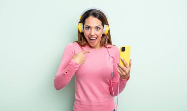 Şok olmuş ve ağzı açık, kendini işaret eden güzel İspanyol bir kadın. akıllı telefon ve kulaklık kavramı