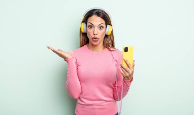 Güzel İspanyol bir kadın şaşırmış ve şok olmuş görünüyor. Çenesi düşmüş ve elinde bir nesne tutuyor. akıllı telefon ve kulaklık kavramı