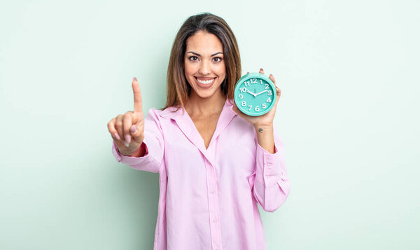 pretty hispanic woman smiling and looking friendly, showing number one. alarm clock concept