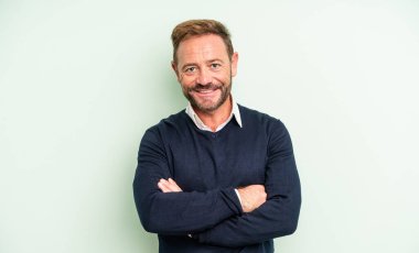 Orta yaşlı yakışıklı adam kameraya çapraz kollarıyla gülümsüyor ve mutlu, kendinden emin, tatmin olmuş bir ifade, yan görüş.