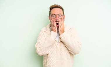 Orta yaşlı yakışıklı adam şok olmuş ve korkmuş hissediyor, ağzı açık ve elleri yanaklarında korkmuş görünüyor.
