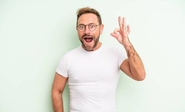 Orta yaşlı yakışıklı adam kendini başarılı ve tatmin olmuş hissediyor, ağzı açık gülümsüyor, elle işaret veriyor.