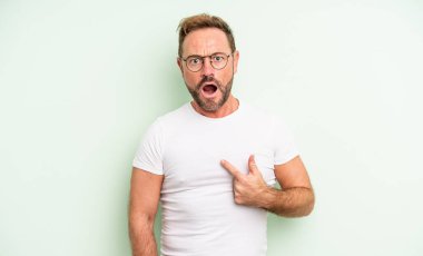 Orta yaşlı yakışıklı bir adam şok olmuş ve şaşırmış görünüyor ağzı açık, kendini işaret ediyor.
