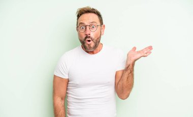 Orta yaşlı yakışıklı bir adam şaşırmış ve şoka uğramış görünüyor, çenesi düşmüş, bir objeyi elinde açık bir elle tutuyor.