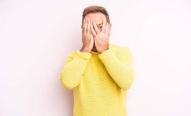 Orta yaşlı yakışıklı adam elleriyle yüzünü kapatıyor, parmaklarının arasından şaşkın bir ifadeyle yan tarafa bakıyor.