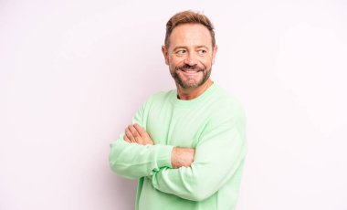 Orta yaşlı yakışıklı adam neşeyle gülümsüyor, mutlu, tatmin olmuş ve rahatlamış hissediyor, kollarını kavuşturup yan tarafa bakıyor.