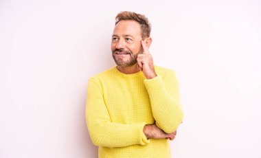 Orta yaşlı yakışıklı adam mutlu bir şekilde gülümsüyor ve hayal kuruyor ya da şüphe ediyor, yan tarafa bakıyor.