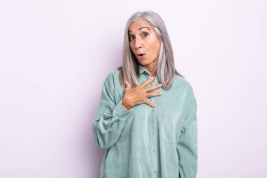 Orta yaşlı beyaz saçlı kadın şaşkın, şaşkın ve şaşırmış hissediyor, elleri göğsünde ve ağzı açık, kim olduğumu söylüyor, ben?
