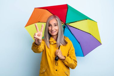 Orta yaşlı, gri saçlı, gülümseyen ve mutlu görünen bir kadın zafer ya da huzur işareti yapıyor. şemsiye ve yağmur kavramı