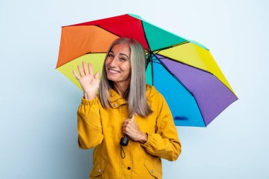 Orta yaşlı gri saçlı kadın mutlu bir şekilde gülümsüyor, el sallıyor, sizi selamlıyor ve selamlıyor. şemsiye ve yağmur kavramı