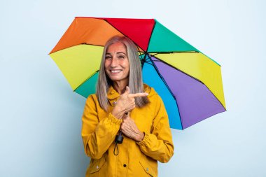 Orta yaşlı, gri saçlı, neşeli gülümseyen, mutlu hisseden ve bir tarafı işaret eden bir kadın. şemsiye ve yağmur kavramı