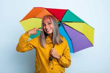 Orta yaşlı, beyaz saçlı, kendine güvenen bir kadın kendi gülümsemesini işaret ediyor. şemsiye ve yağmur kavramı