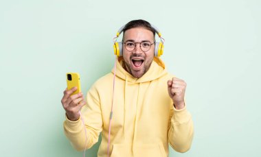 Genç yakışıklı adam şok olmuş hissediyor, gülüyor ve başarıyı kutluyor. kulaklıklar ve akıllı telefon konsepti