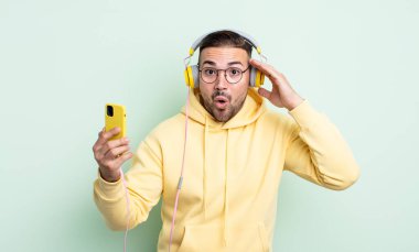 Genç yakışıklı adam mutlu, şaşkın ve şaşırmış görünüyor. kulaklıklar ve akıllı telefon konsepti