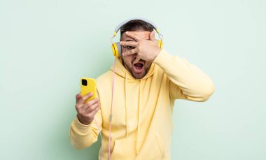 Genç, yakışıklı adam şok olmuş, korkmuş ya da korkmuş görünüyor. kulaklıklar ve akıllı telefon konsepti