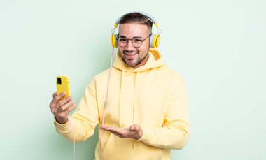 Genç yakışıklı adam neşeyle gülümsüyor, mutlu hissediyor ve bir konsept gösteriyor. kulaklıklar ve akıllı telefon konsepti