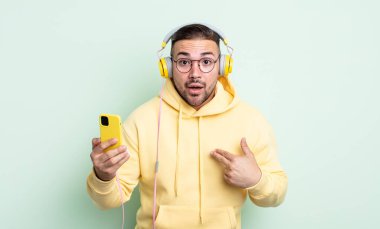 Şok olmuş ve ağzı açık, kendini işaret eden genç ve yakışıklı bir adam. kulaklıklar ve akıllı telefon konsepti