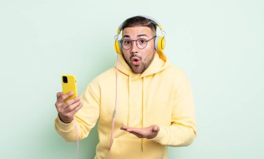 Genç, yakışıklı adam şaşırmış ve şok olmuş görünüyor. Çenesi düşmüş, elinde bir nesne var. kulaklıklar ve akıllı telefon konsepti